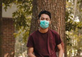 Man In Maroon Crew Neck T-Shirt With Face Mask Standing Beside Brown Tree During Daytime
