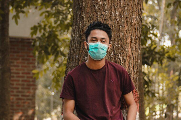 Man In Maroon Crew Neck T-Shirt With Face Mask Standing Beside Brown Tree During Daytime