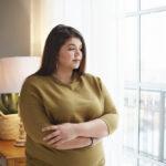 people lifestyle body positivity concept beautiful young brunette female with extra pounds standing by window home crossing arms her chest having thoughtful pensive look pondering