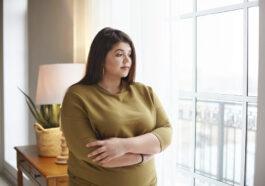 people lifestyle body positivity concept beautiful young brunette female with extra pounds standing by window home crossing arms her chest having thoughtful pensive look pondering